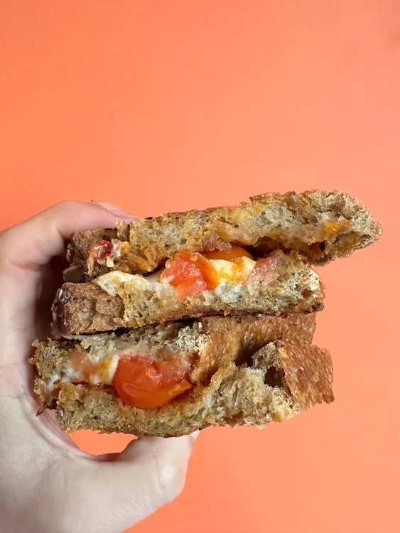 Een hand die een tosti met kaas, sambal en tomaat vast houdt voor een oranje achtergrond