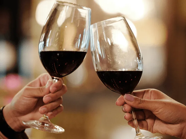 Twee handen die allebei een glas rode wijn vast houden. De wijnglazen proosten met elkaar. 