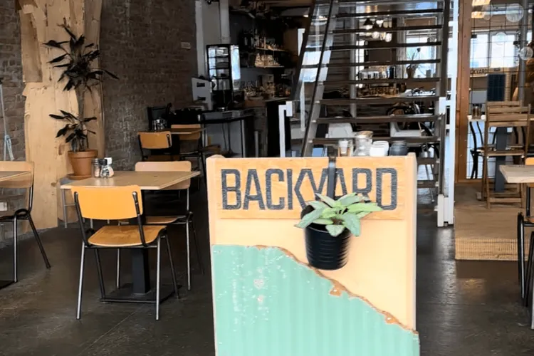 De entree van Backyard restaurant. Je ziet een trap die omhoog gaat en een houten tafel met houten stoelen. Ook zie je een houten bord met de naam "backyard" en een plantje dat daar aan hangt. 