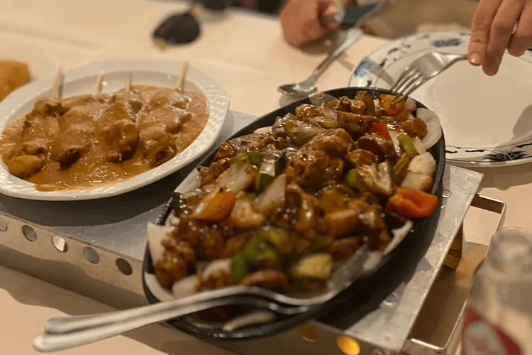 Een warmhoud plaatje met daarop twee gerechten. Op het linker bord zie je een bruine saus met stokjes, sate. Rechts zie je een gietijzeren plaatje met een vleesvervanger en groente zoals paprika en ui. Hierin ligt bestek om het mee op te scheppen.