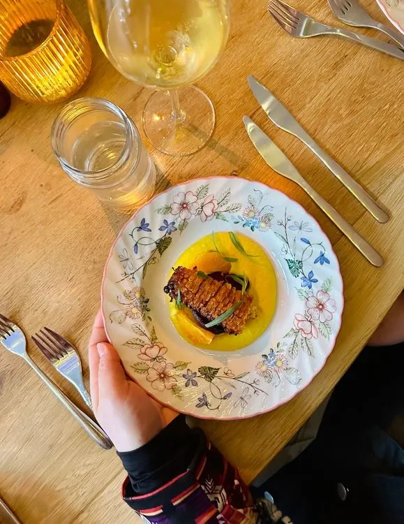 Een hand houdt een wit bord vast met erop een gele crème, een mais kolf en ingelegde bloedsinaasappel