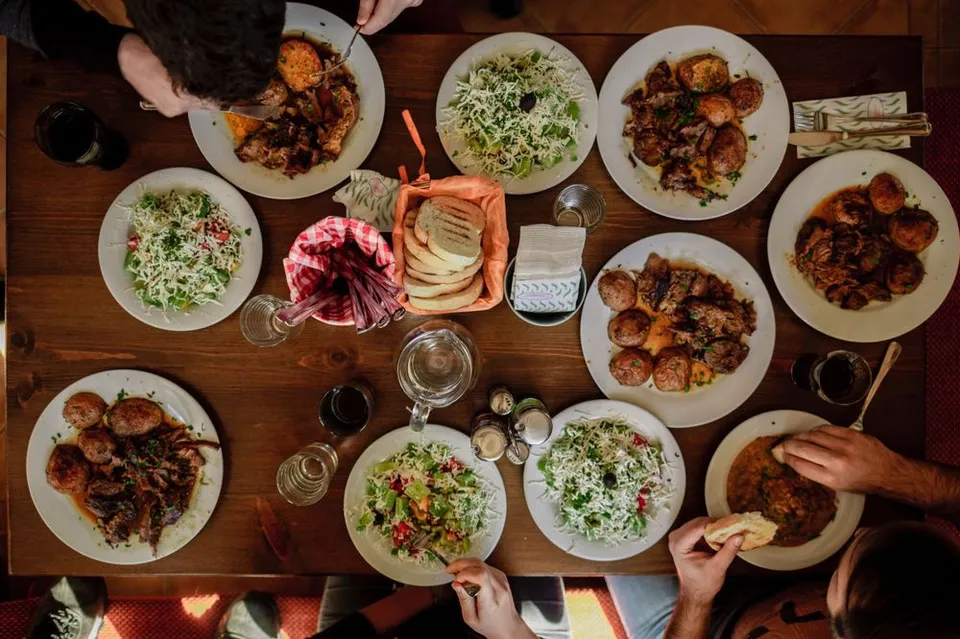 Een tafel van boven vol met allerlei kleurrijke gerechten. Ook zie je drie mensen die er om heen zitten en met hun handen gerechten aanraken. 